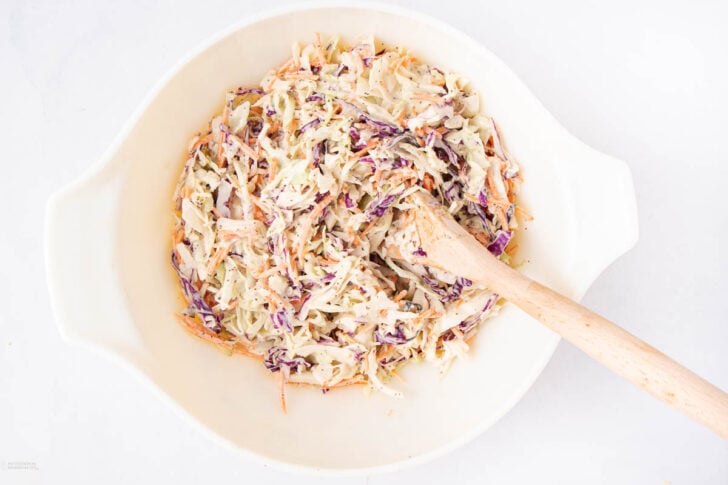 A white bowl filled with creamy coleslaw, containing shredded green and purple cabbage and carrots, with a wooden spoon resting inside on a white background.