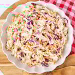 A white scalloped bowl filled with creamy coleslaw made of shredded green and purple cabbage and carrots, resting on a wooden board next to a red and white checkered cloth.
