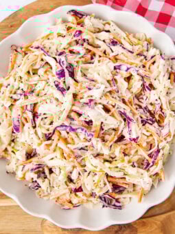 A white scalloped bowl filled with creamy coleslaw made of shredded green and purple cabbage and carrots, resting on a wooden board next to a red and white checkered cloth.