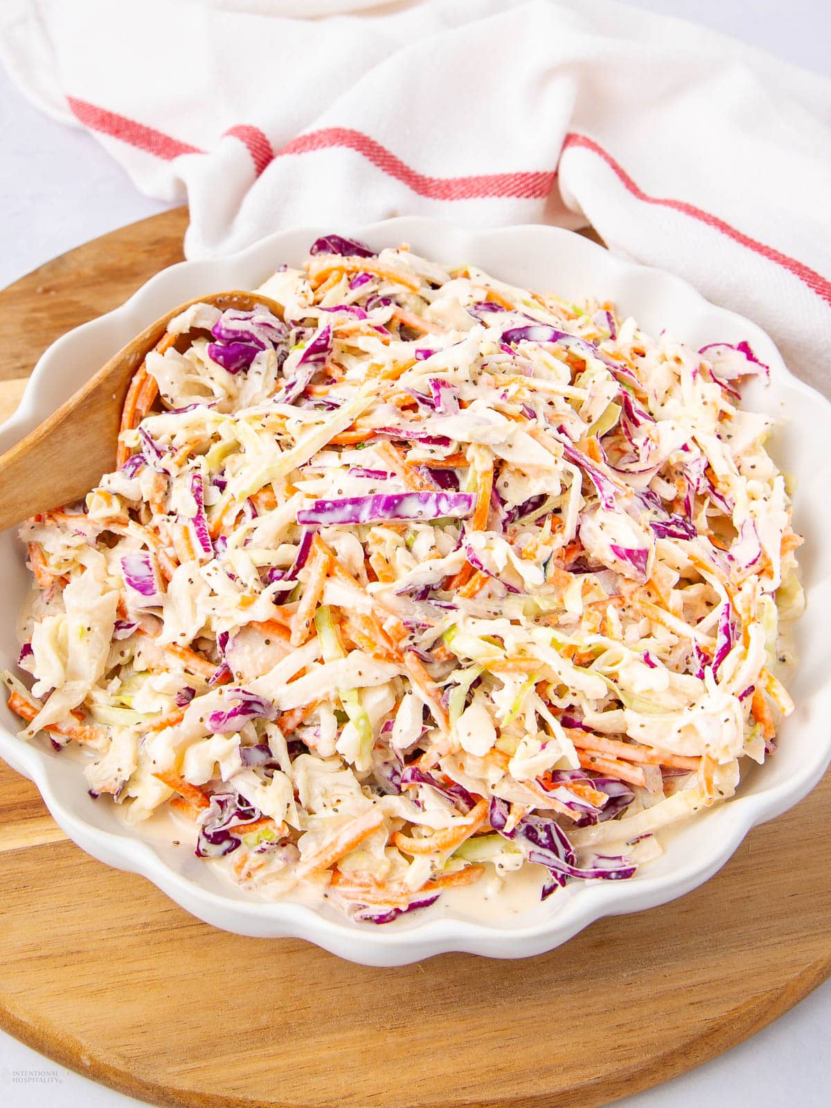A bowl of creamy coleslaw with shredded green cabbage, purple cabbage, and carrots sits on a wooden board, with a wooden spoon and a white cloth with a red stripe in the background.