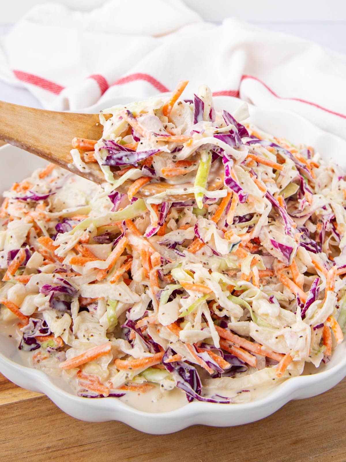 A close-up of creamy coleslaw with shredded green and purple cabbage and carrots, being served with a wooden spoon from a white scalloped bowl. A white cloth is in the background.