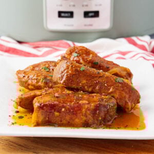 A white plate with several pieces of saucy, seasoned boneless pork ribs garnished with chopped herbs. A slow cooker and a red-and-white striped towel are visible in the background.