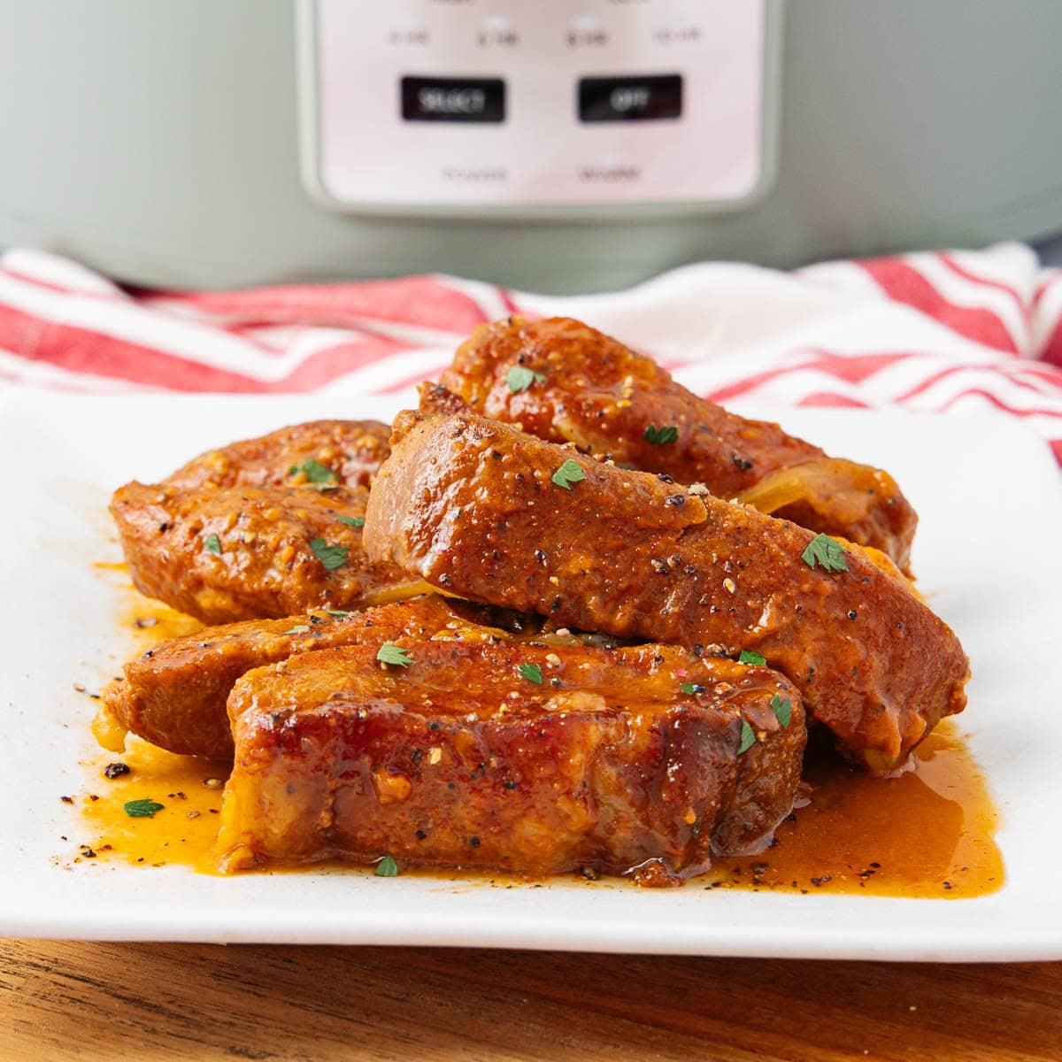 A white plate with several pieces of saucy, seasoned boneless pork ribs garnished with chopped herbs. A slow cooker and a red-and-white striped towel are visible in the background.