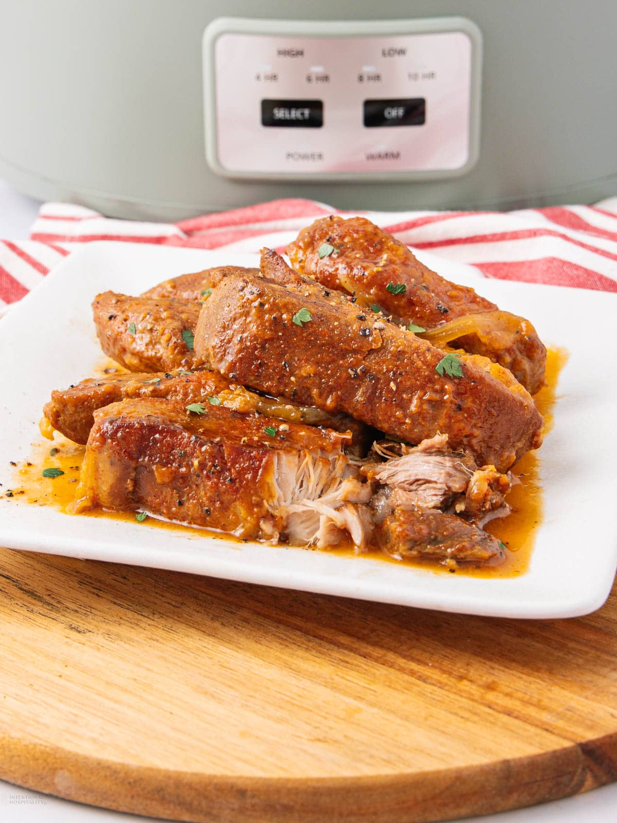 A plate of shredded, seasoned pork in sauce sits on a white dish atop a wooden board, with a slow cooker and a red-striped towel in the background.