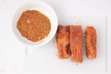 Three pieces of raw meat coated with dry seasoning are placed next to a small white bowl filled with extra seasoning mix, all on a white cutting board against a white background.