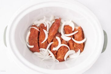 Raw, seasoned chicken breasts and sliced onions placed in a white slow cooker, ready to be cooked. The chicken is coated with spices, and the onions are scattered on top and around the meat.