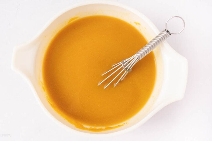 A white mixing bowl filled with a smooth orange-brown sauce or batter, with a metal whisk resting inside, on a white background.