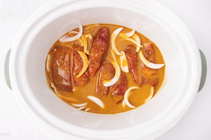 Sliced sausages and raw onion wedges in a creamy orange sauce inside a white slow cooker, viewed from above.