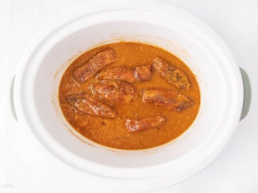 Chicken drumsticks cooked in a rich, orange-brown sauce inside a white slow cooker, viewed from above on a white background.