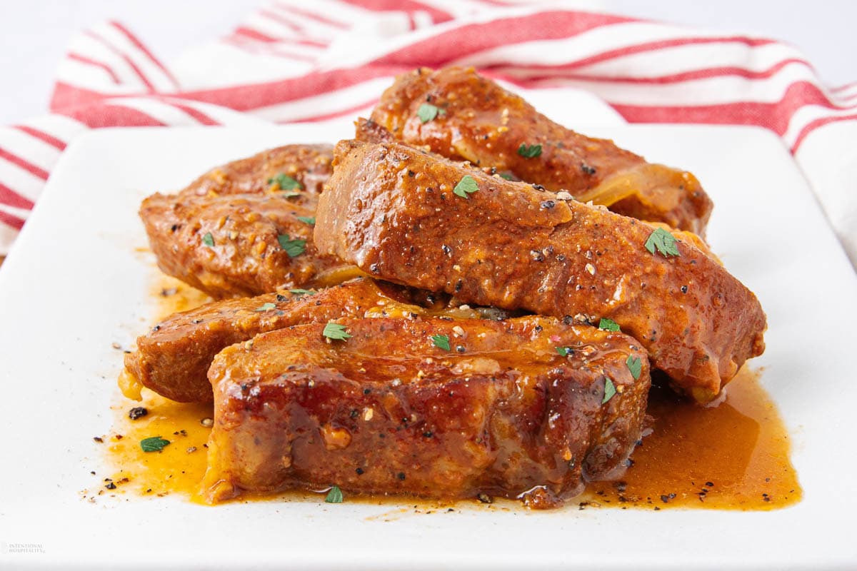 Four pieces of cooked pork ribs covered in a rich, reddish-brown sauce, garnished with chopped herbs, served on a white plate with a red-and-white striped cloth in the background.