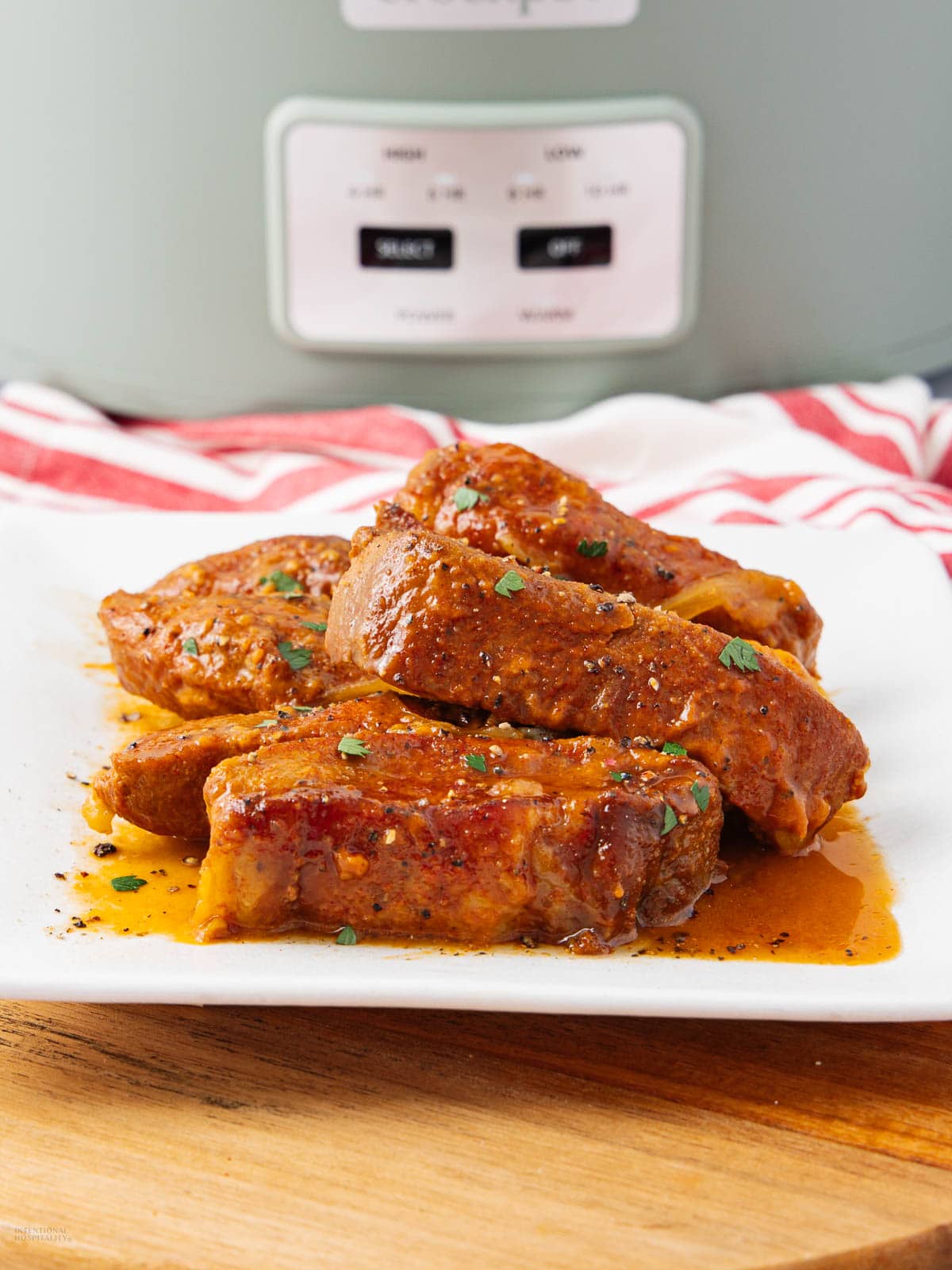 Four saucy, seasoned boneless pork ribs are served on a white plate, garnished with chopped herbs, with a slow cooker and a red-and-white striped towel in the background.