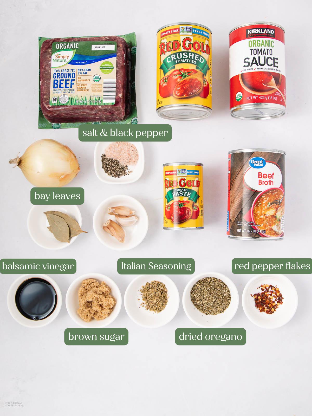 Flat lay of spaghetti sauce ingredients: ground beef, crushed tomatoes, tomato sauce, beef broth, onion, garlic, bay leaves, balsamic vinegar, Italian seasoning, brown sugar, dried oregano, red pepper flakes, salt, and black pepper.
