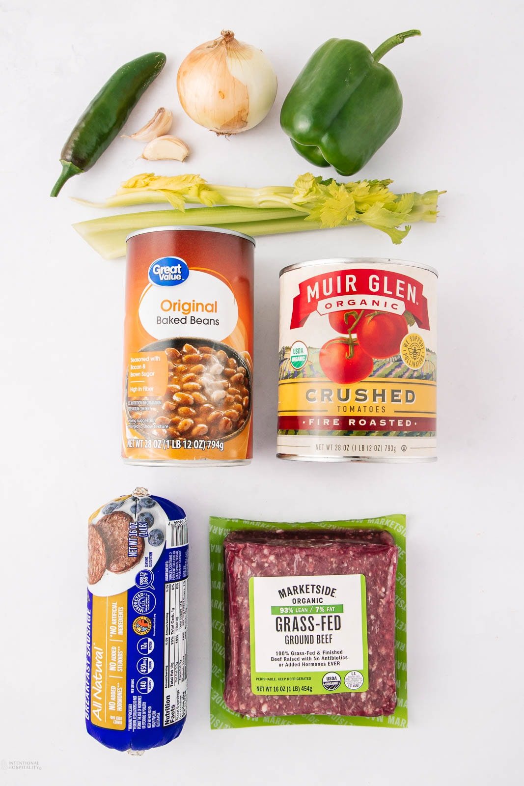 Fresh jalapeño, onion, garlic, green bell pepper, celery, a can of baked beans, a can of crushed tomatoes, a roll of ground turkey, and a package of grass-fed ground beef arranged on a white background.