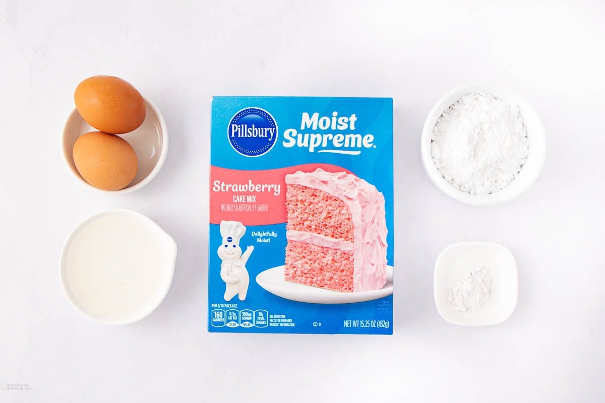A box of Pillsbury Moist Supreme Strawberry Cake Mix is surrounded by two eggs, a bowl of milk, a bowl of flour, and a bowl of baking powder on a white surface.