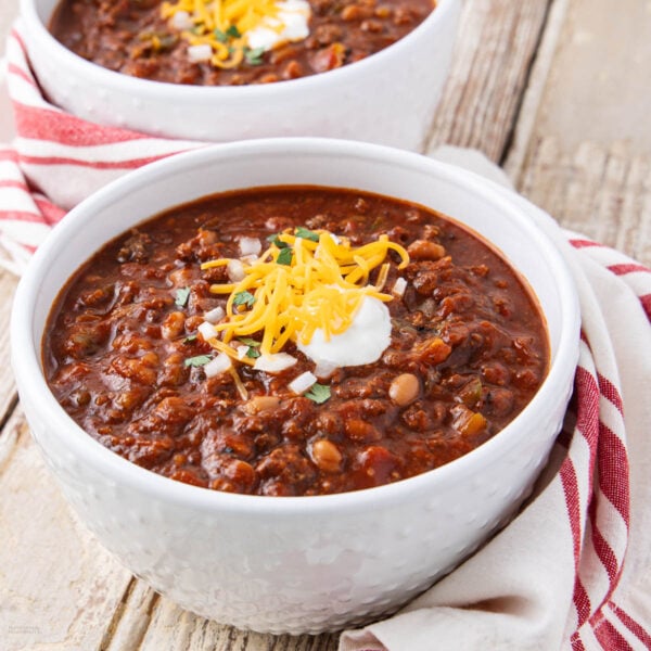 A white bowl filled with hearty chili topped with shredded cheese, sour cream, and chopped green onions, placed on a rustic wooden table with a red and white striped napkin.