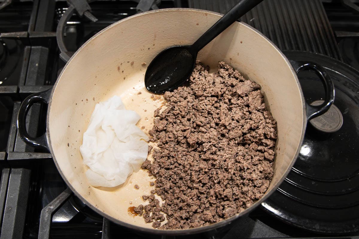 A pot on a stove contains cooked ground beef on one side and a wad of white paper towels used for absorbing grease on the other side, with a black spoon resting inside the pot.