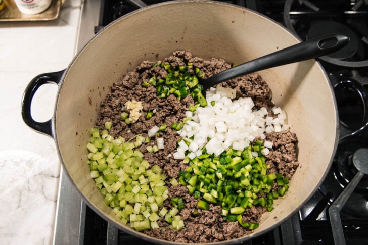 A pot on a stove containing cooked ground beef, chopped onions, celery, green bell pepper, jalapeño, and minced garlic, with a black spoon resting inside.