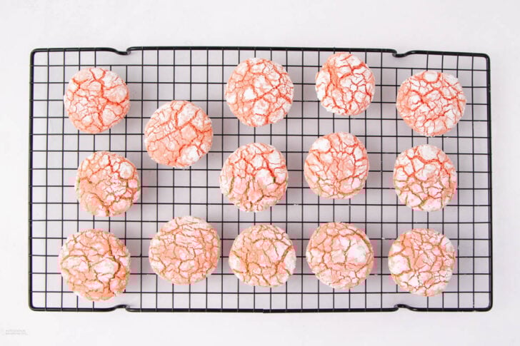 Rows of pink crinkle cookies dusted with powdered sugar cooling on a black wire rack against a white background.