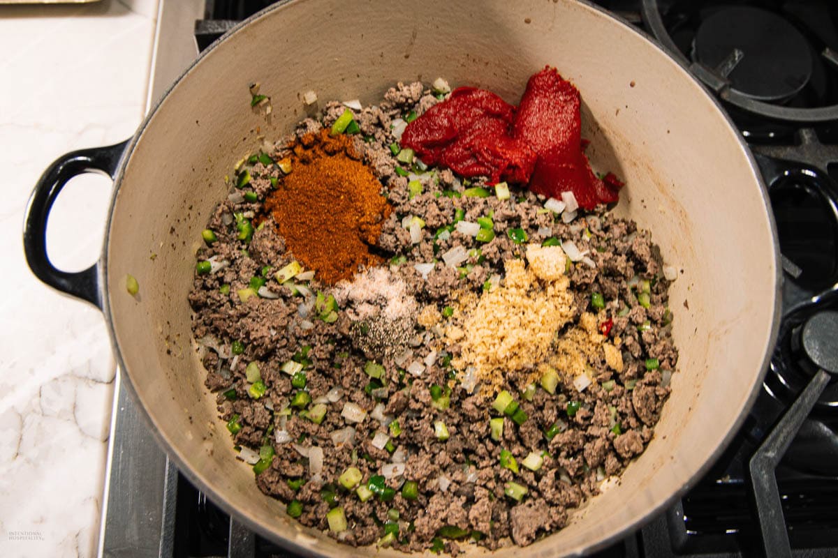 Ground beef, chopped green peppers, and onions are cooking in a pot. On top are mounds of tomato paste, chili powder, and seasonings, not yet mixed in. The pot sits on a stovetop.