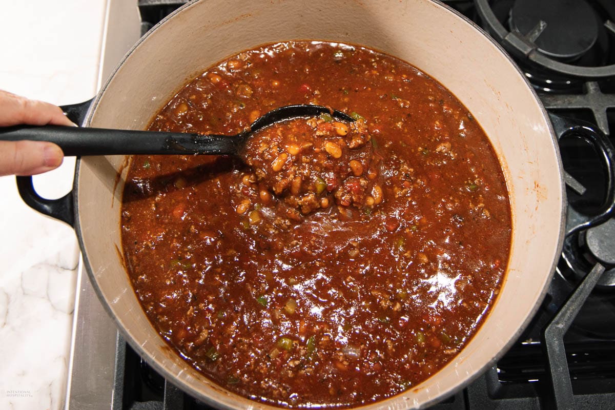A person is holding a ladle of chunky chili above a pot on a stove. The chili is red and filled with beans, ground meat, and vegetables.