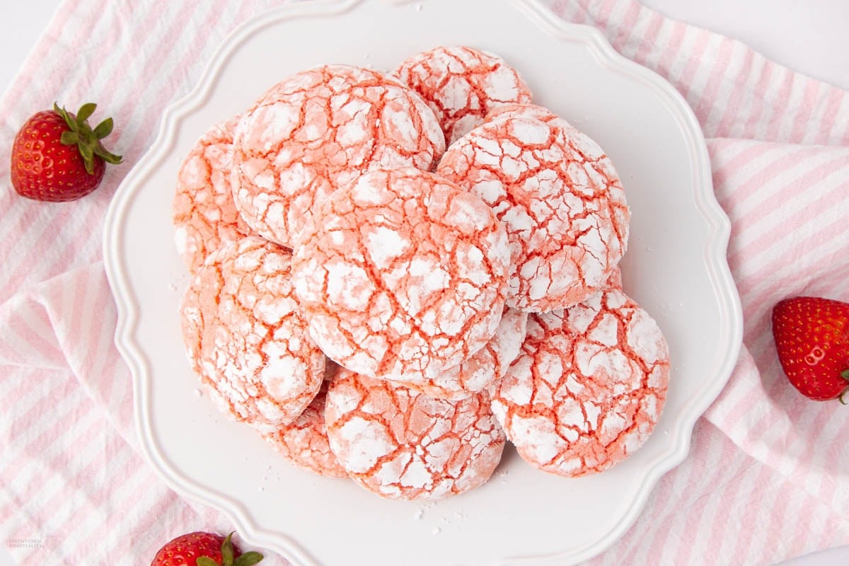 A plate of pink strawberry crinkle cookies dusted with powdered sugar, arranged on a white plate. Fresh strawberries are placed nearby on a pink and white striped cloth.