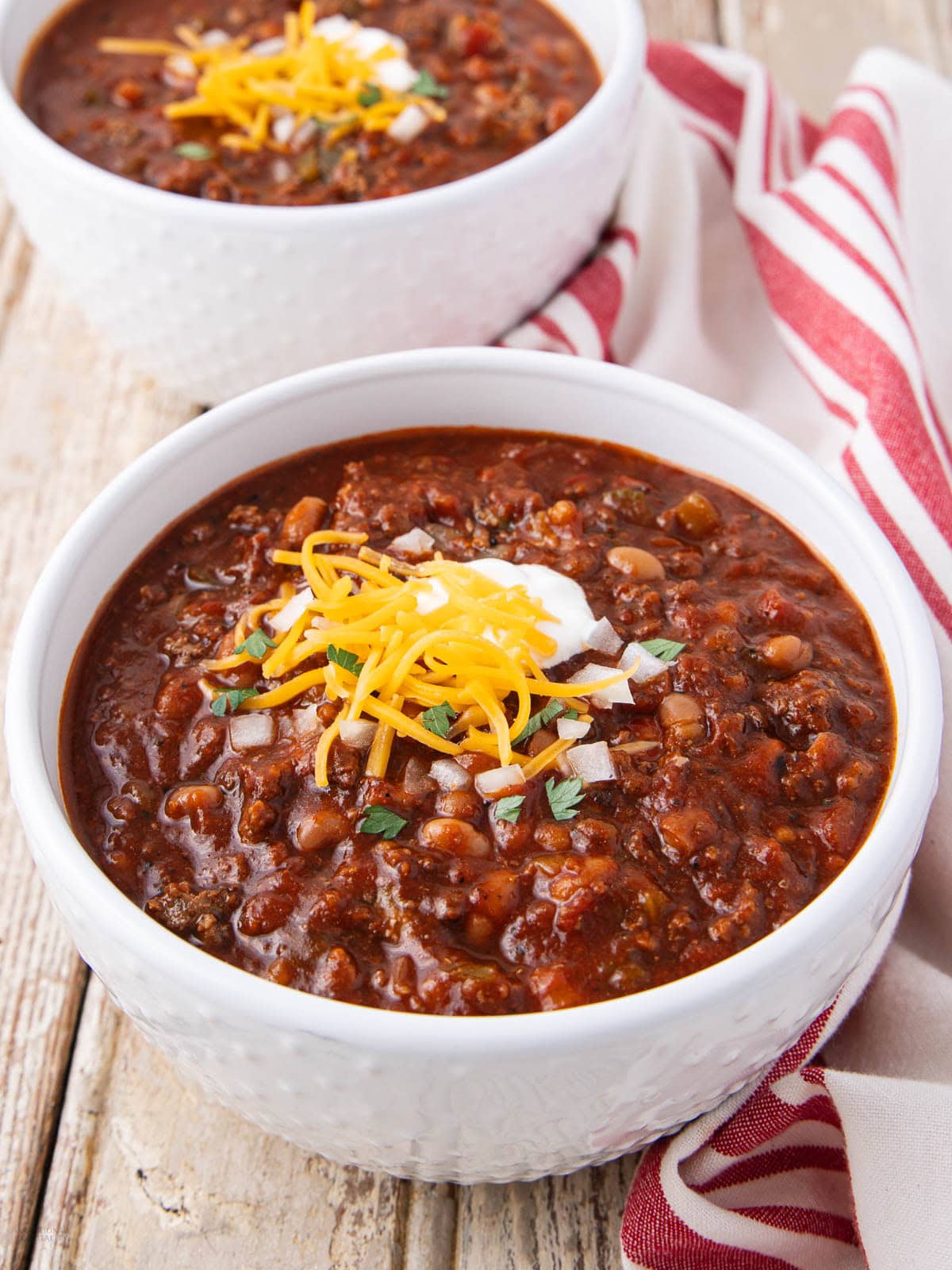 Two white bowls of chili topped with shredded cheese, sour cream, and chopped onions, set on a rustic wooden table with a red and white striped cloth nearby.