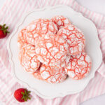A plate of strawberry crinkle cookies dusted with powdered sugar, arranged on a white decorative plate. Fresh strawberries sit nearby on a pink and white striped cloth.