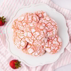 A plate of strawberry crinkle cookies dusted with powdered sugar, arranged on a white decorative plate. Fresh strawberries sit nearby on a pink and white striped cloth.