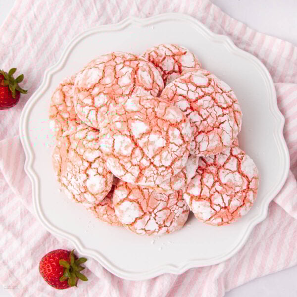 A plate of strawberry crinkle cookies dusted with powdered sugar, arranged on a white decorative plate. Fresh strawberries sit nearby on a pink and white striped cloth.