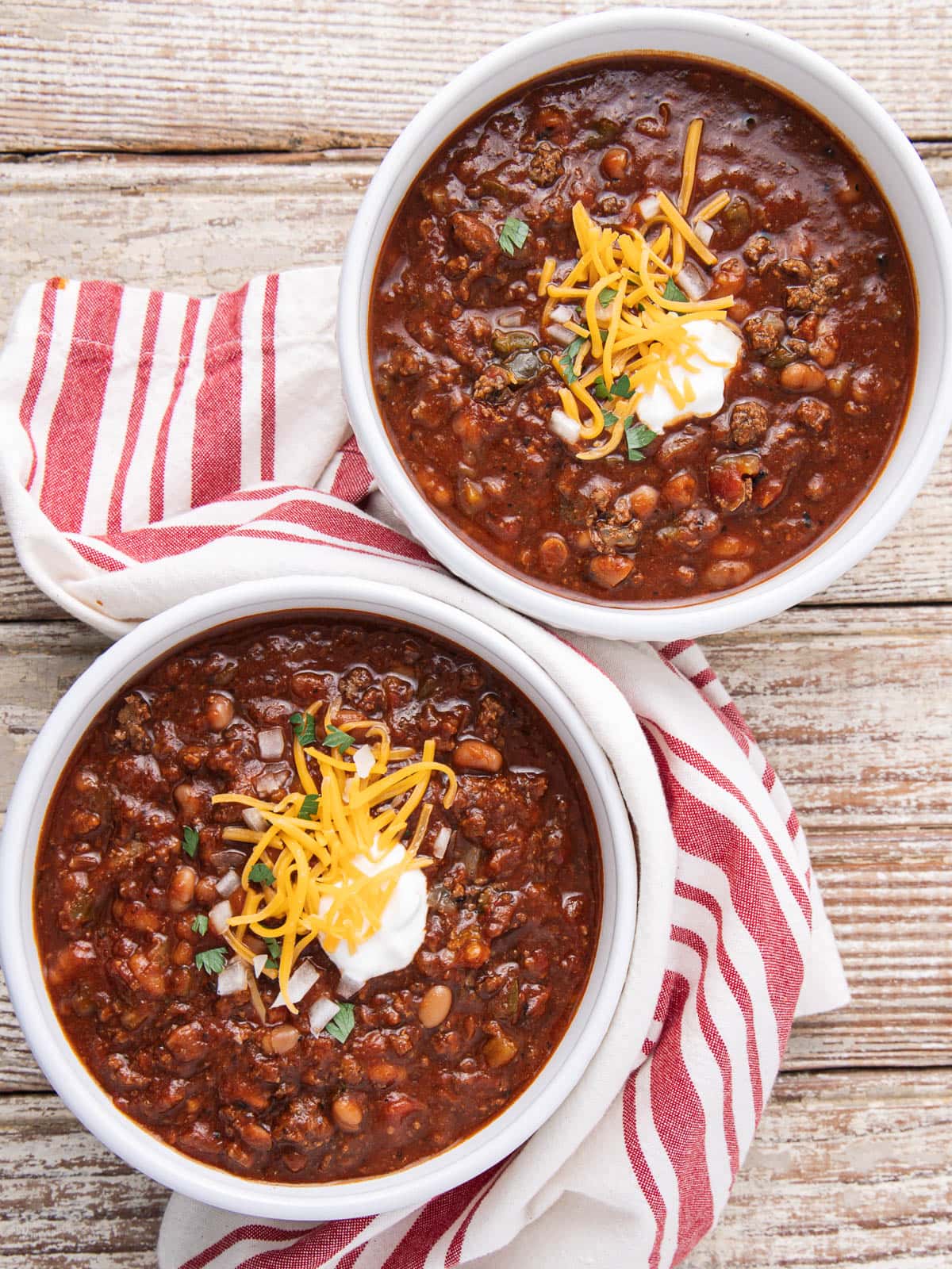 Two bowls of chili topped with shredded cheddar cheese and a dollop of sour cream, placed on a rustic wooden surface with red and white striped napkins underneath each bowl.