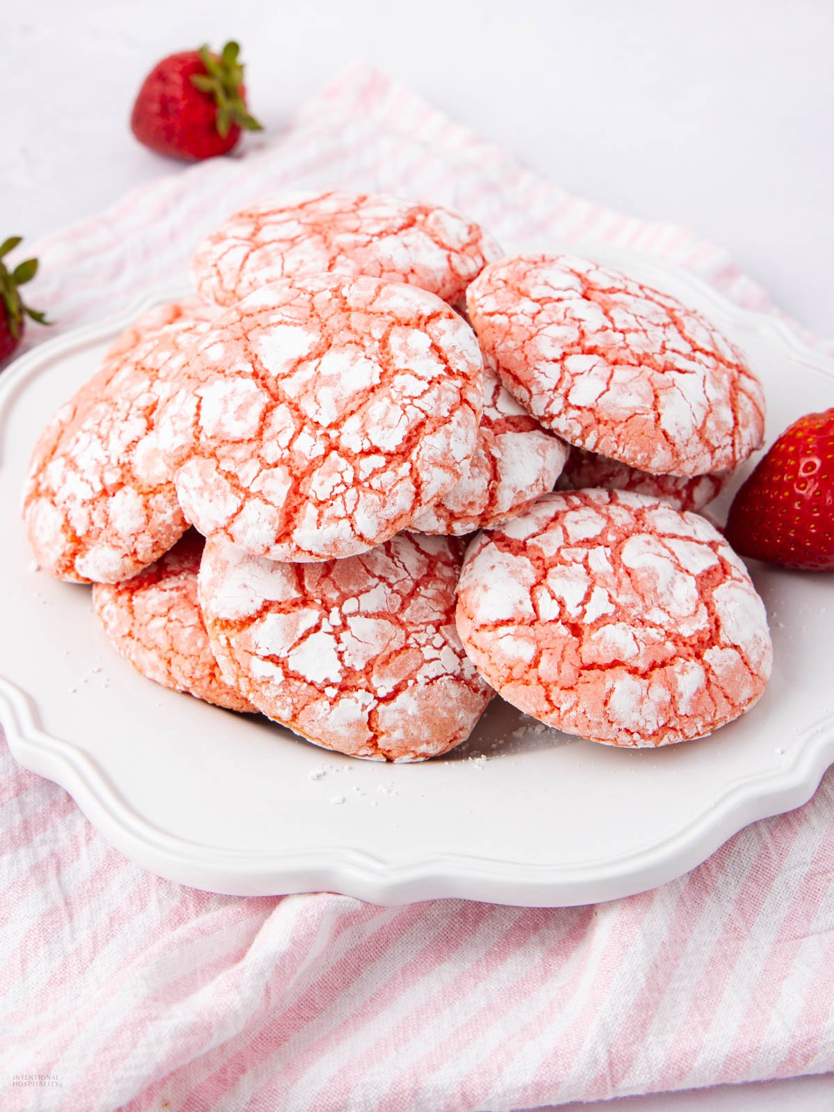 A plate of pink crinkle cookies dusted with powdered sugar sits on a white plate, with a few fresh strawberries and a pink-striped cloth in the background.