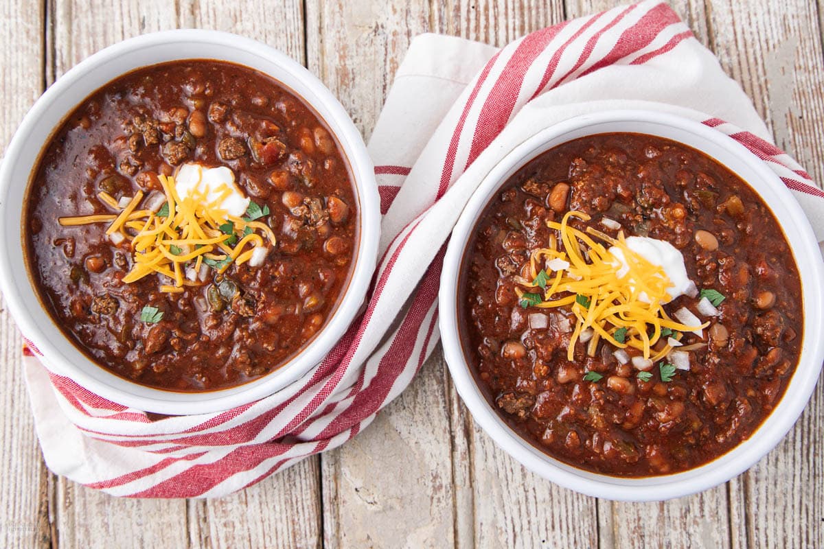 Two bowls of chili topped with shredded cheese, sour cream, and chopped herbs, set on a rustic wooden table with red and white striped towels underneath each bowl.