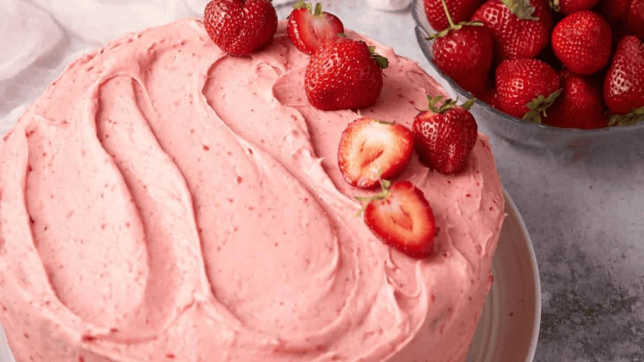 A round cake covered in smooth pink strawberry frosting, topped with fresh strawberries. A bowl of whole strawberries sits nearby on a light surface.