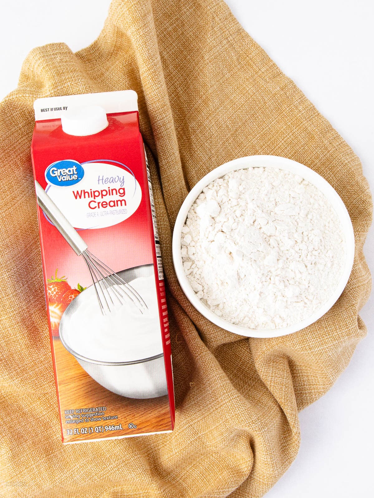 A carton of Great Value heavy whipping cream next to a white bowl filled with flour, both placed on a textured tan cloth.