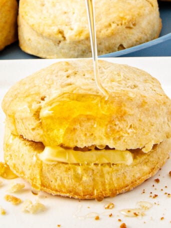 A golden biscuit sliced open with a pat of butter inside, being drizzled with honey. Crumbs are scattered on the white plate, and more biscuits are visible in the background.