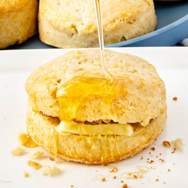 A golden biscuit sliced open with a pat of butter inside, being drizzled with honey. Crumbs are scattered on the white plate, and more biscuits are visible in the background.