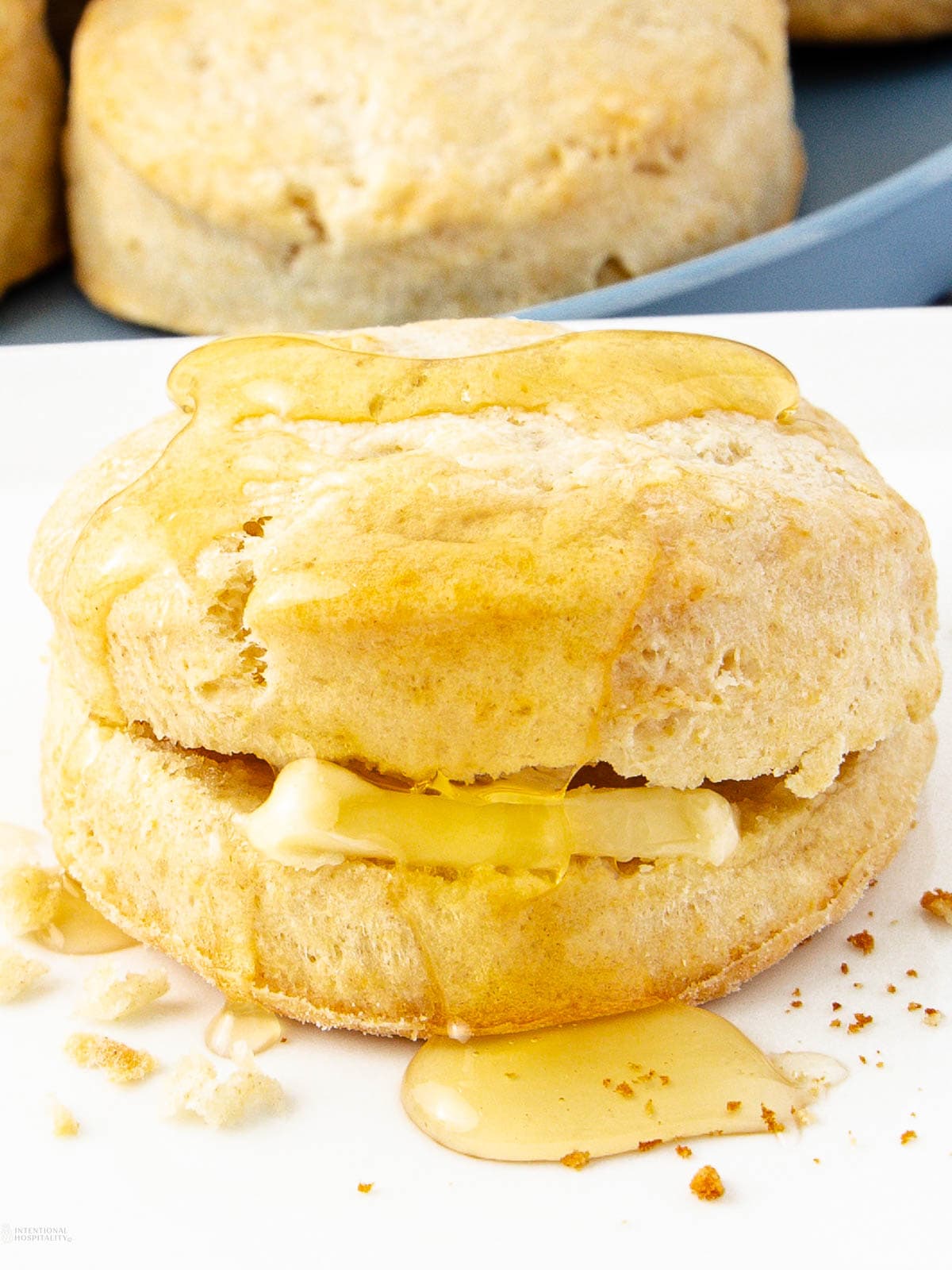 A golden, flaky biscuit is split open with a pat of melting butter inside, drizzled generously with honey. Crumbs and honey drops are scattered on the white plate beneath the biscuit.