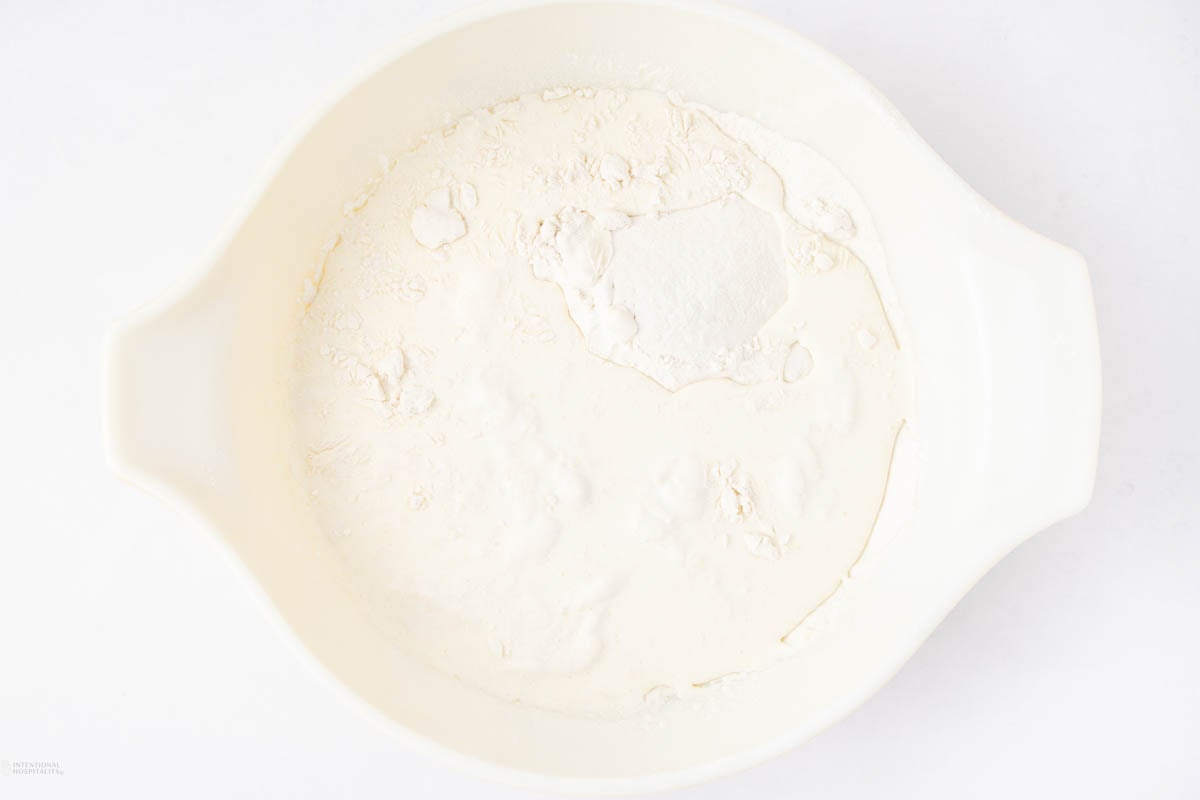 A white mixing bowl filled with a mound of white flour and a small amount of sugar, viewed from above on a white background.