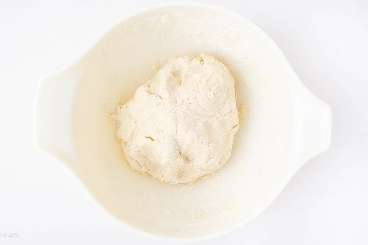A ball of raw dough sits in the center of a white mixing bowl on a white background.