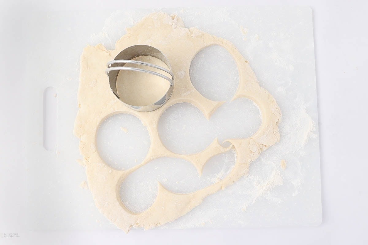 A round metal cookie cutter is pressed into rolled-out dough on a white cutting board, creating circular shapes for baking.