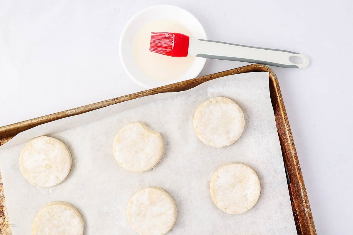 A parchment-lined baking sheet holds six unbaked round biscuits. A red pastry brush rests on a white bowl with liquid, ready for brushing the biscuits.