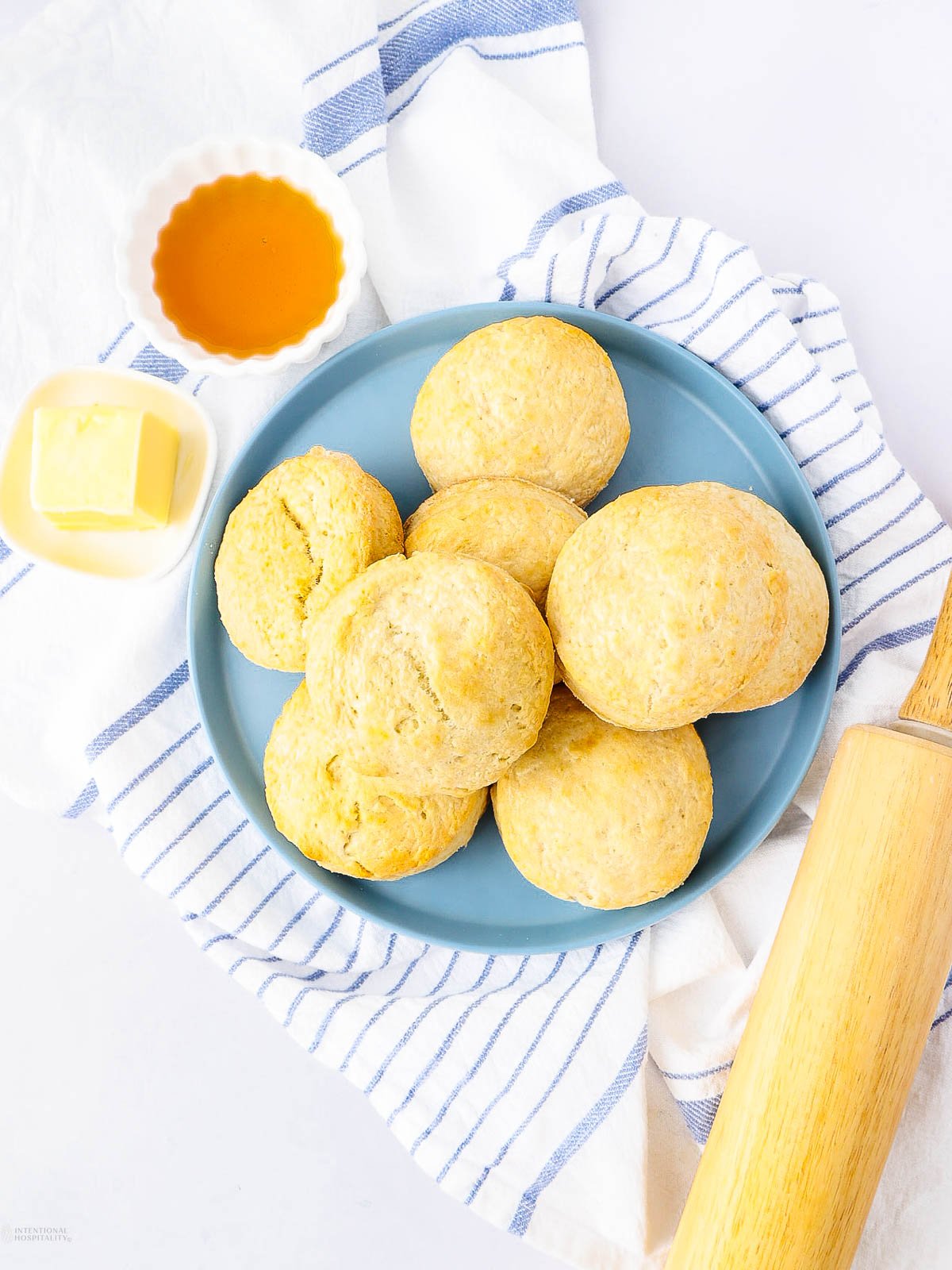 A blue plate with seven biscuits sits on a white and blue striped towel. Nearby are a rolling pin, a small dish of honey, and a dish with a pat of butter.