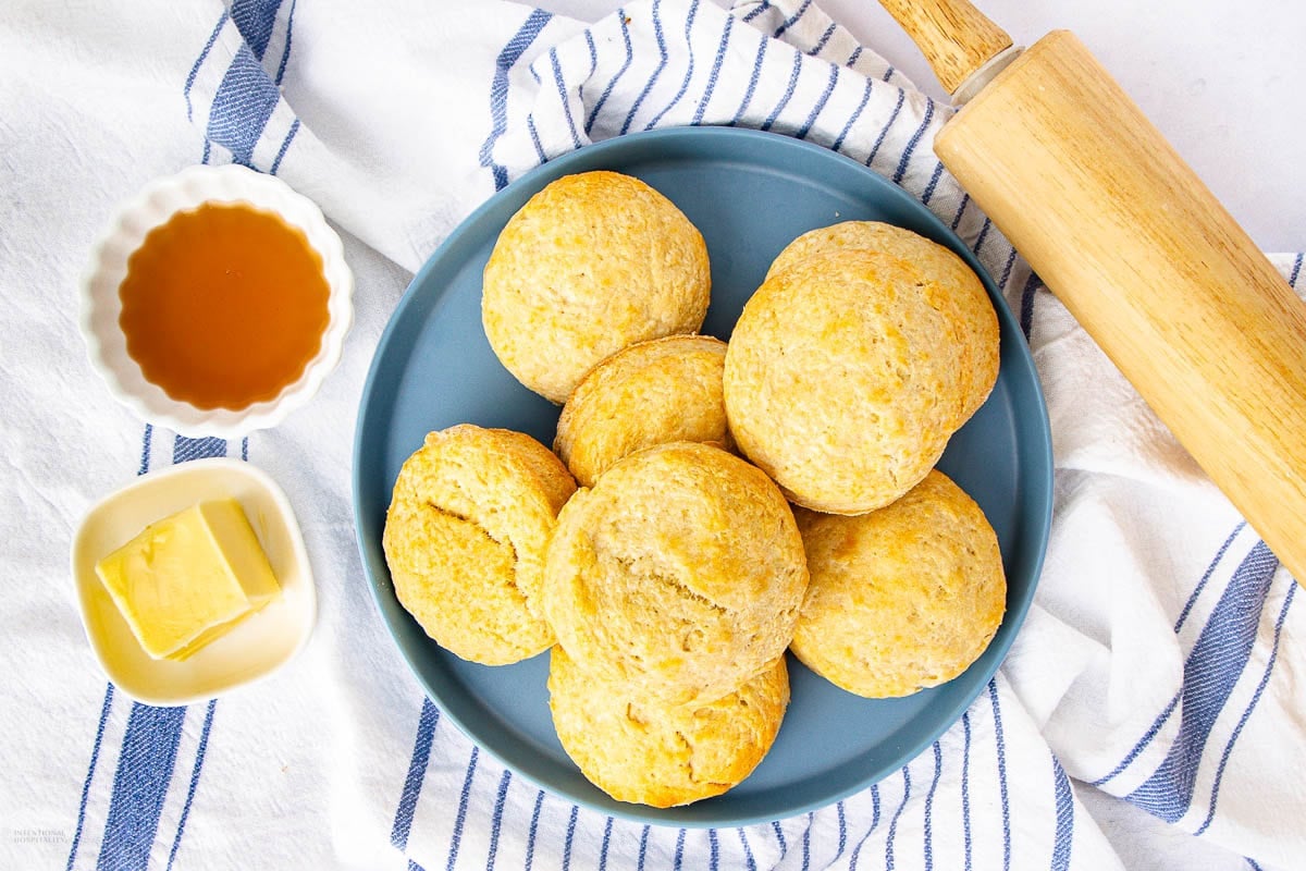 A blue plate with six 2 ingredient biscuits sits on a striped towel. Nearby are a small dish of butter, a dish of honey, and a rolling pin.