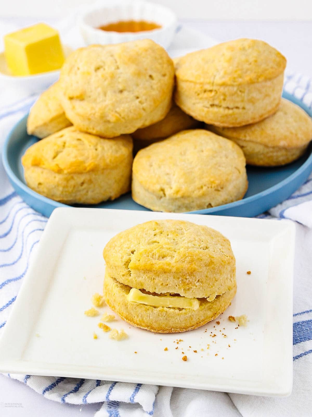 A plate with several golden brown 2 ingredient biscuits stacked on it, with a single biscuit split and filled with butter on a white plate in the foreground, and a dish of butter in the background.