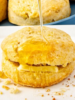 A golden 2 ingredient biscuit split open with butter inside, drizzled with honey, sits on a white plate with crumbs nearby.