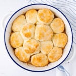 A white round baking dish filled with golden-brown biscuits, arranged closely together, sits on a white surface next to a blue-striped kitchen towel.