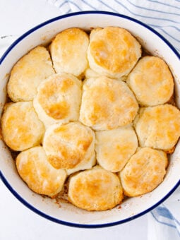 A white round baking dish filled with golden-brown biscuits, arranged closely together, sits on a white surface next to a blue-striped kitchen towel.