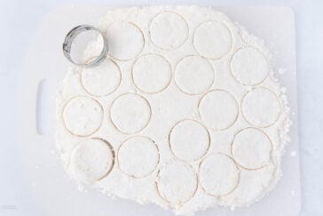 A sheet of biscuit dough with round shapes cut out using a metal biscuit cutter, resting on a floured surface.