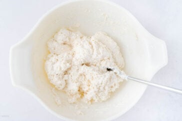 A white mixing bowl containing a sticky, lumpy dough mixture with a metal spoon partially submerged in it, placed on a white surface.