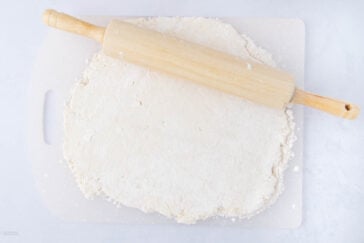 A wooden rolling pin rests on top of a slab of dough being rolled out on a white cutting board, with flour scattered around.